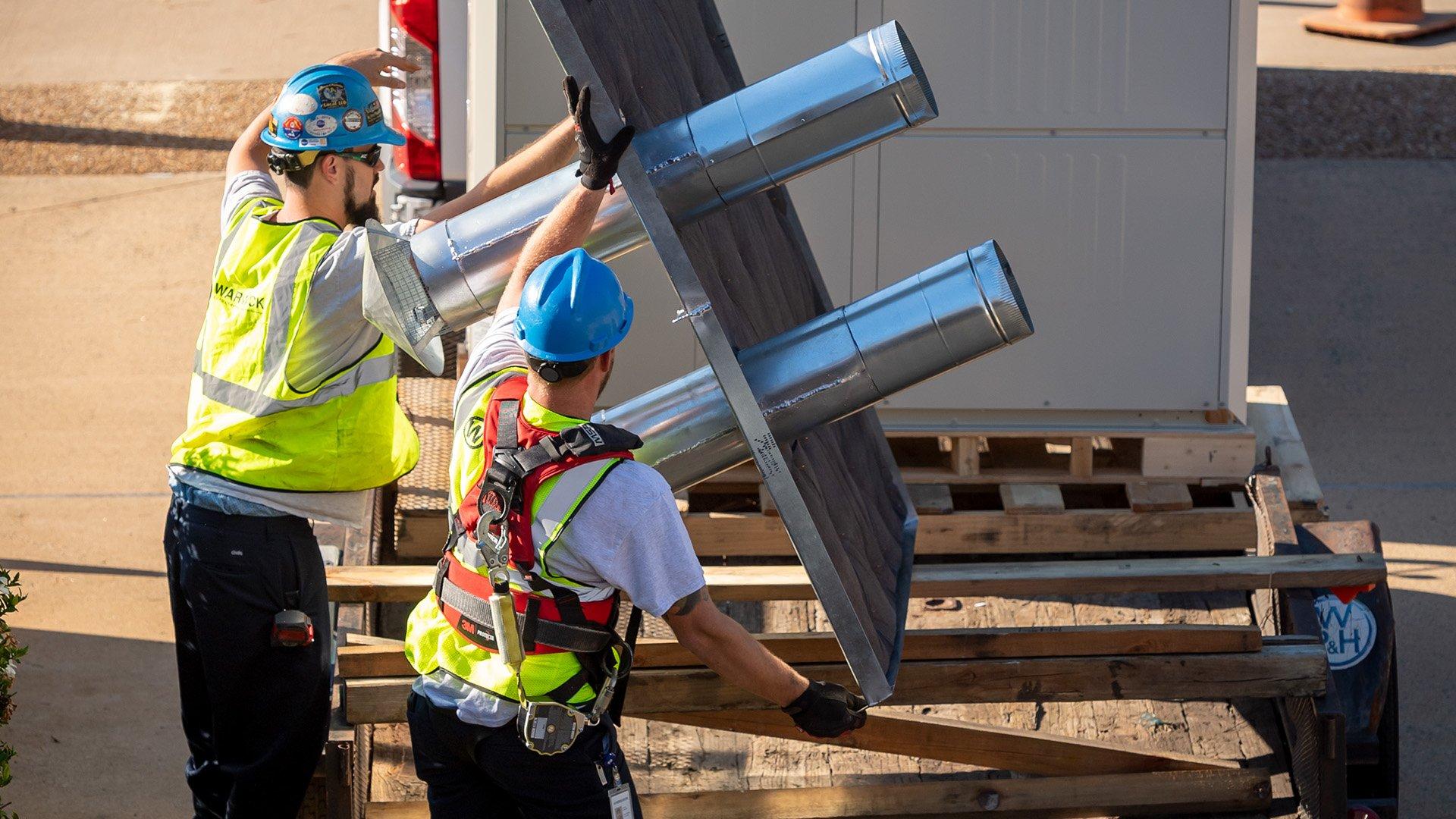 Two workers in safety vests and blue hardhats lift piping from palette. 